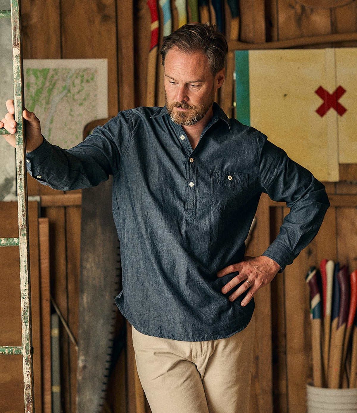 A man in the Best Made Co. Indigo Chambray Popover and beige pants stands indoors, leaning on a ladder, with woodworking tools and colorful artwork in the background.