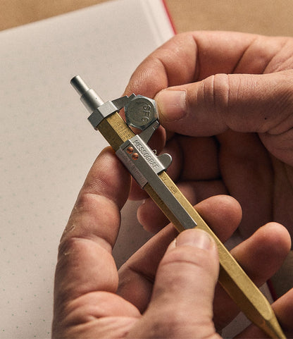 A person adjusts the Best Made Co. Brass Messograf Pen, featuring a metal clip and eraser, holding it above a dotted notebook—a display of precision and ingenuity that embodies German craftsmanship.