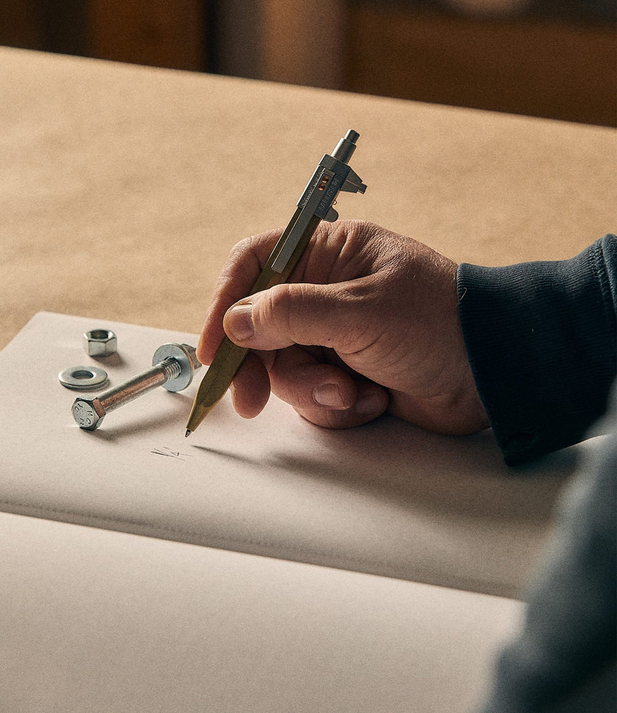 A hand writes in a notebook using the Best Made Co. Brass Messograf Pen, a fine example of German craftsmanship, while a bolt, nut, and washer rest nearby on the open pages.