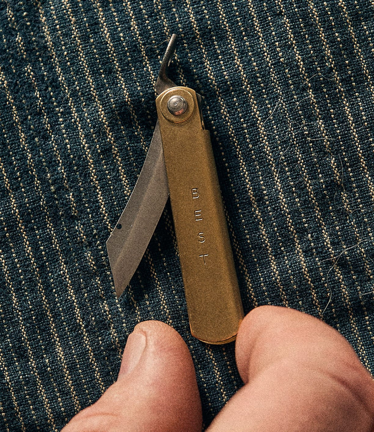 A Mini Brass Higo Knife by Best Made Co. rests on blue striped fabric, near a person’s fingertips.