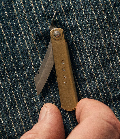 A Mini Brass Higo Knife by Best Made Co. rests on blue striped fabric, near a person’s fingertips.