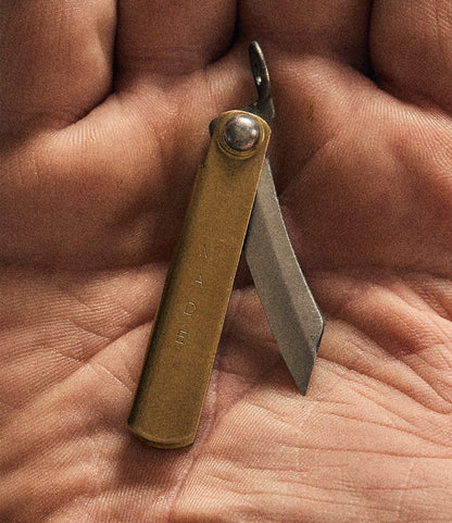 A Mini Brass Higo Knife by Best Made Co. with a partially opened blade rests on the palm of a hand. The brass handle is engraved with “MADE,” showcasing the craftsmanship of this handmade knife.