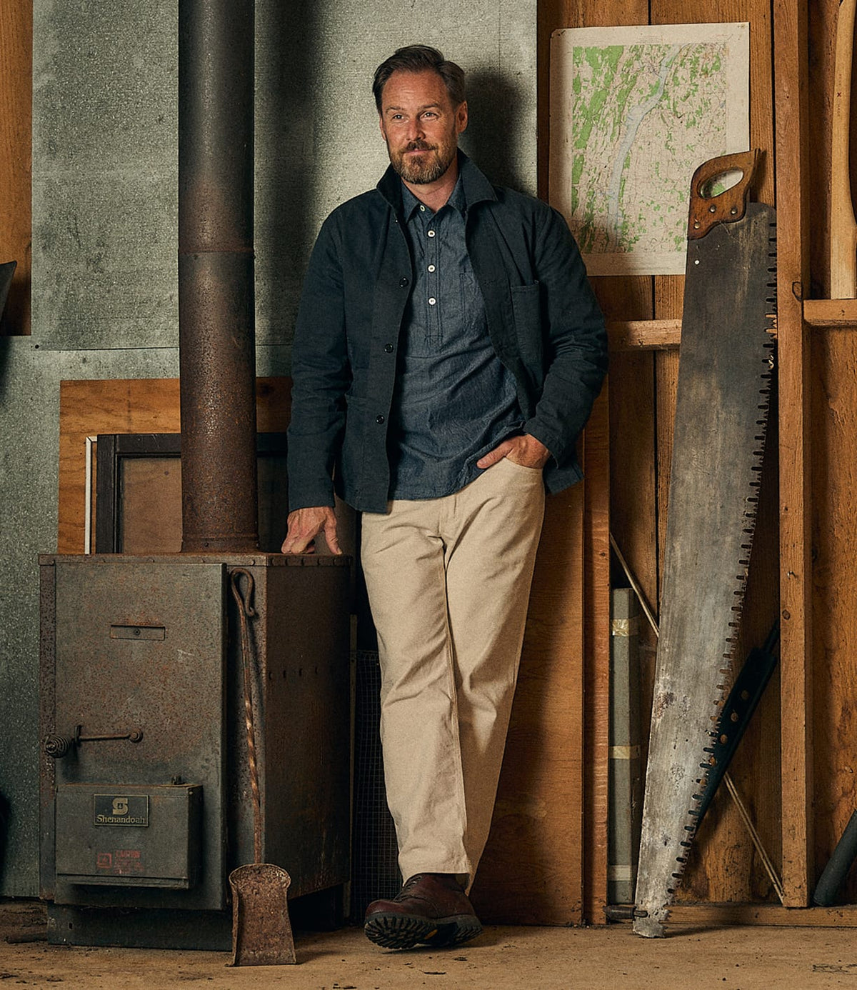 Wearing the Best Made Co. Service Jacket Special Edition Navy and light pants, a man leans against a wood stove in a rustic workshop, with a large saw and an axe nearby. A framed map hangs on the wooden wall behind him.