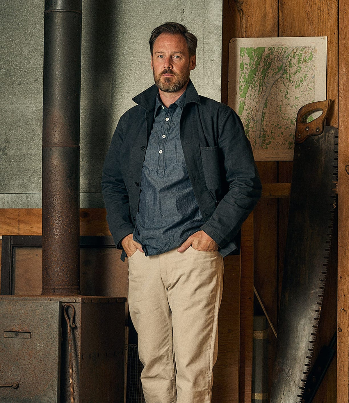 A bearded man stands indoors beside a wood stove, wearing the Best Made Co. Service Jacket Special Edition Navy over a blue shirt and beige pants. Behind him, a large saw and a map hang on the wooden wall.