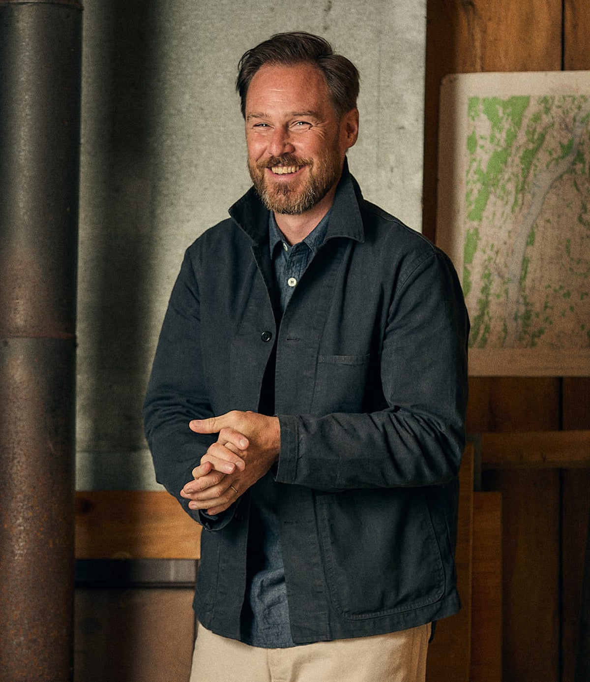 A bearded man smiles while wearing the Best Made Co. Service Jacket Special Edition Navy over a button-up shirt, standing indoors near a metal pipe with a map displayed on a wooden wall in the background.