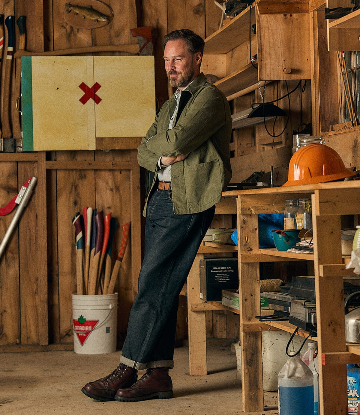 A man with a beard leans against a workbench in a wooden shed, surrounded by tools, shelves, an orange hard hat, and a white bucket. He wears the Best Made Co. Service Jacket Special Edition Olive Drab—an updated take on the classic P-41 Marine Corps style—paired with jeans and boots.