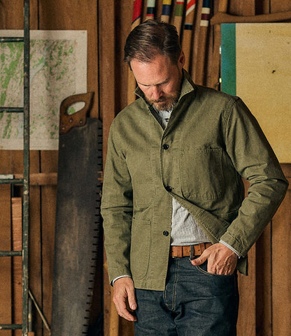 A man wearing the Best Made Co. Service Jacket Special Edition Olive Drab, crafted from Japanese twill, stands in a workshop with wooden walls, tools, and a ladder in the background—an homage to the utilitarian heritage of the classic P-41 Marine Corps style.