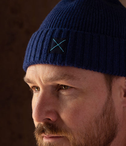 A person wearing a navy blue lambswool Cap of Courage featuring a square patch with an embroidered light blue X.