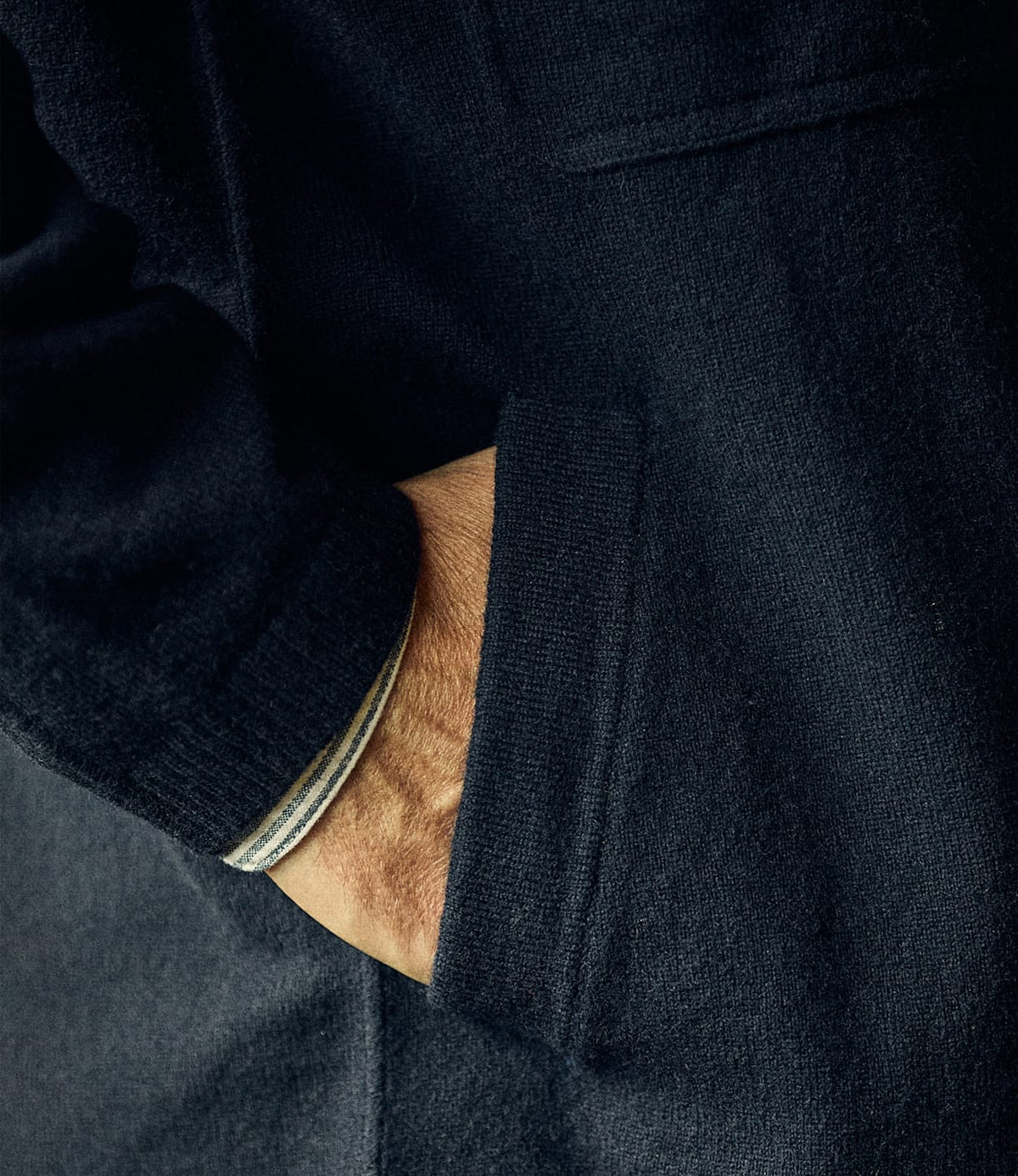 A hand in the pocket of a dark merino wool Knit Lambswool CPO jacket with a striped sleeve cuff visible.