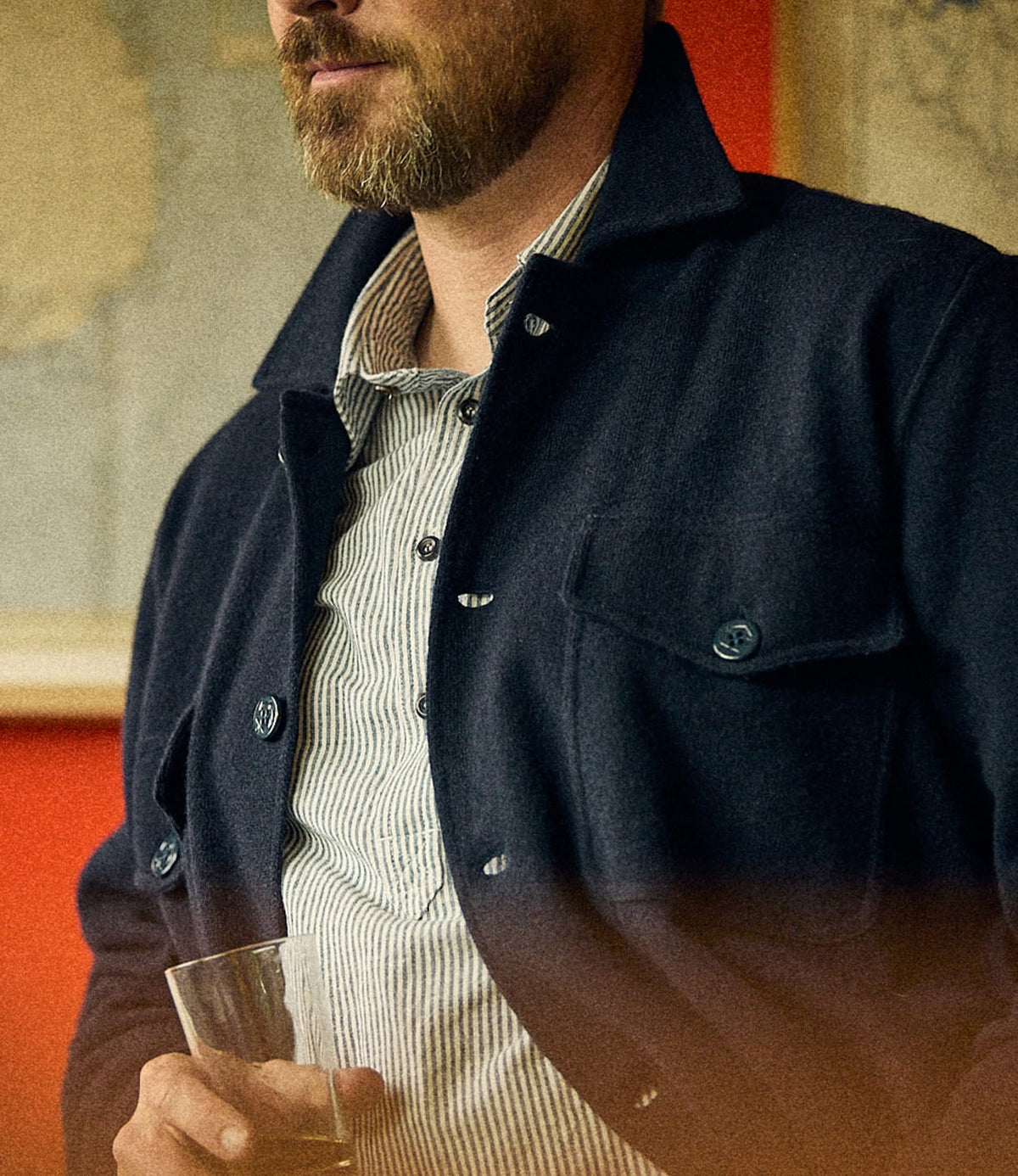A bearded person wears a navy Knit Lambswool CPO over a light-colored, thin-striped collared shirt while holding a glass.