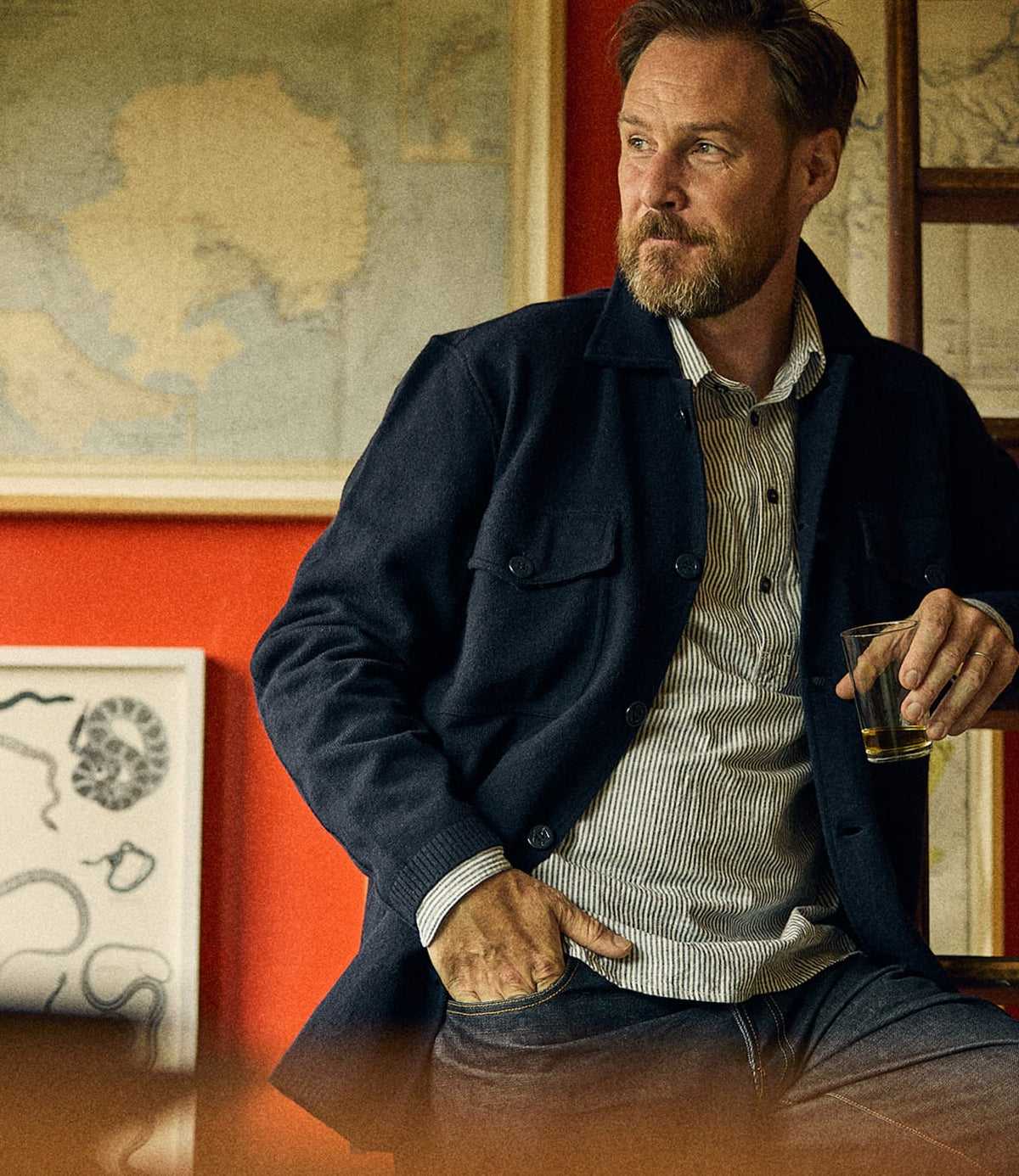 A bearded man wearing a dark navy Knit Lambswool CPO over a striped shirt holds a glass of amber liquid.