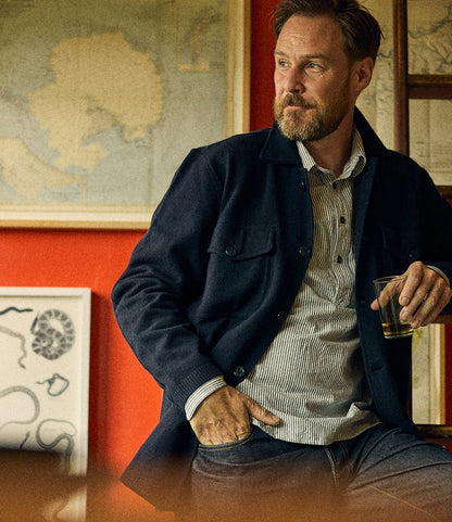 A bearded man wearing a dark navy Knit Lambswool CPO over a striped shirt holds a glass of amber liquid.