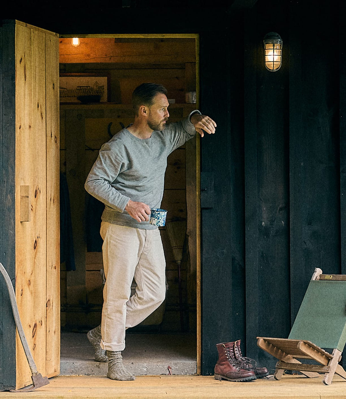 A man in a gray shirt and light pants stands in a wooden doorway, holding a mug and leaning on the doorframe. His relaxed style is complemented by the Best Made Co. SRS Crewneck Heather Gray, custom-knit from soft tanguis cotton French terry. Boots and a chair are on the porch outside.