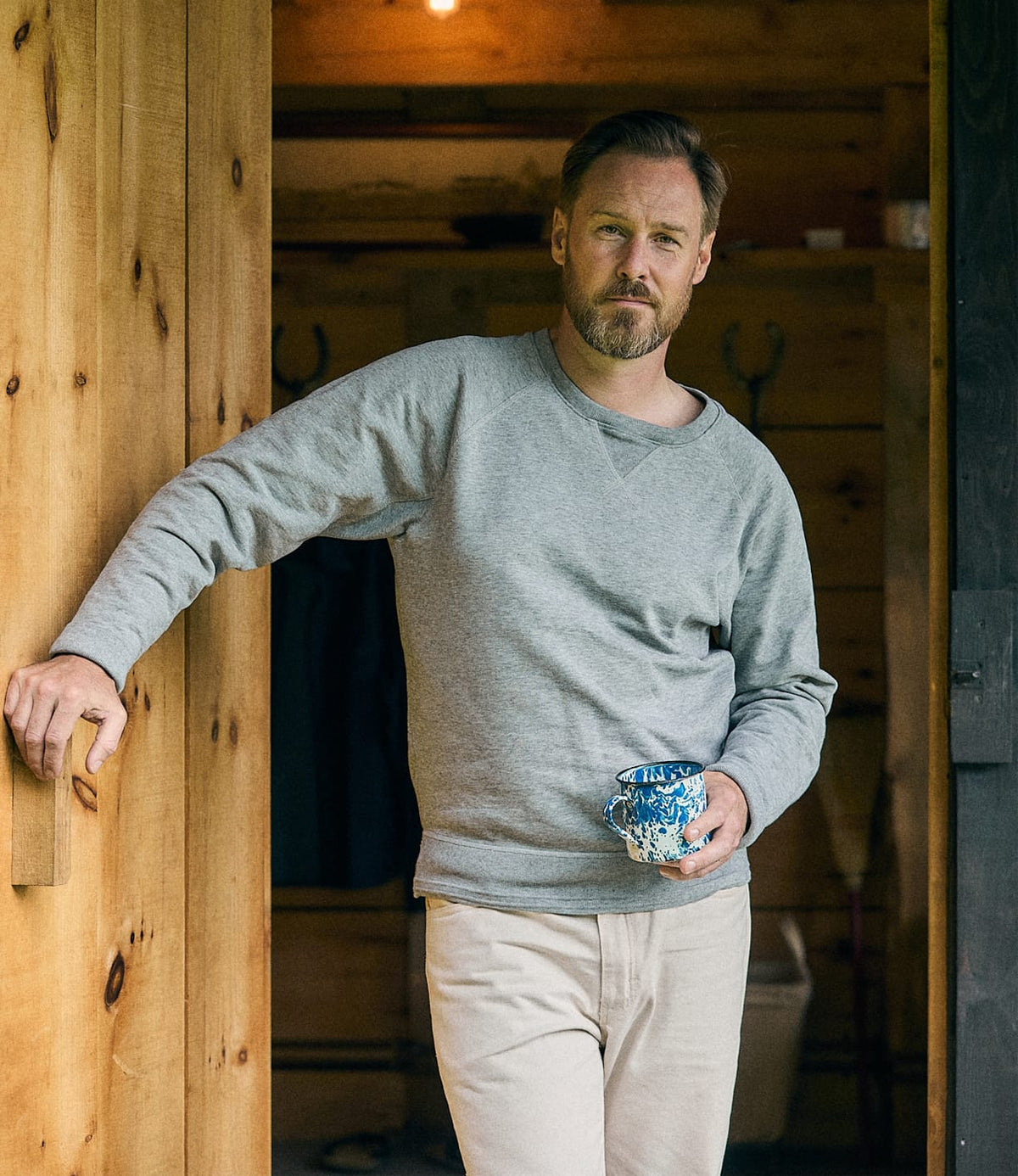 A man with a beard stands in a wooden doorway, wearing the Best Made Co. SRS Crewneck Heather Gray sweatshirt and beige pants, holding a blue patterned mug and looking calmly at the camera.