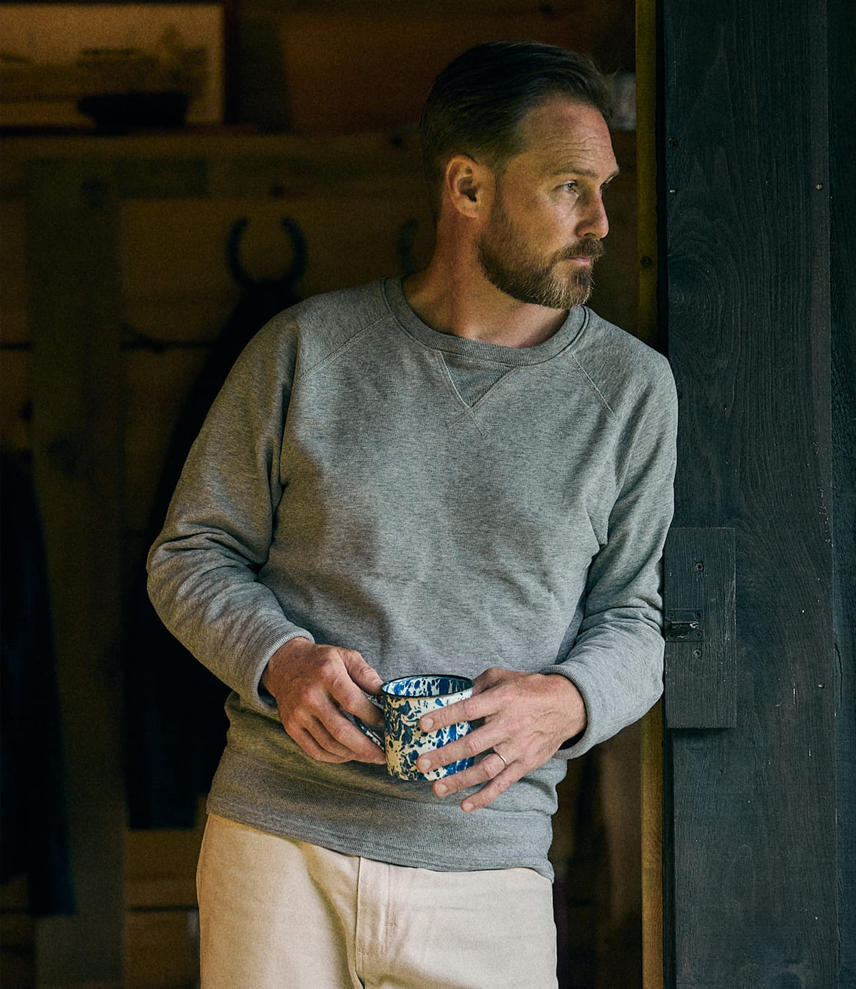 A man with a beard, wearing the Best Made Co. SRS Crewneck Heather Gray—a custom-knit French terry sweatshirt—and light pants, stands by a doorway holding a patterned mug and gazing thoughtfully to his left.