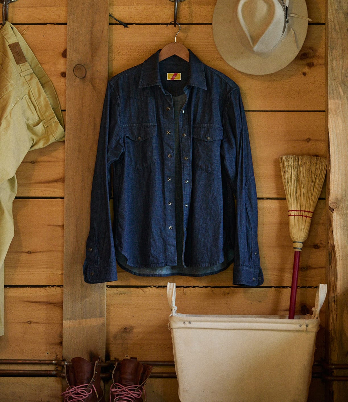A blue Fukuyama Snap-Front Shirt by Best Made Co. in Japanese indigo denim hangs on a wooden wall beside a beige hat, a broom, and khaki pants. Brown boots and a white canvas hamper are placed below.
