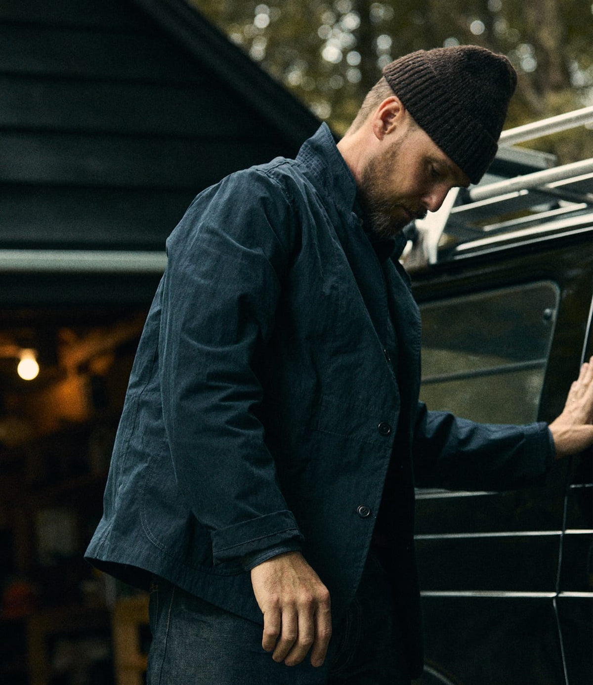 A man wearing the Possibilities Jacket in Navy from Best Made Co. and a brown beanie stands beside a black vehicle, looking down with one hand resting on the car. A building with warm lighting is in the background.