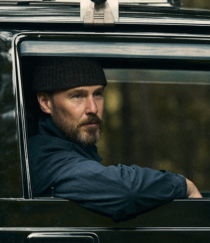 A man with a beard and knit hat, dressed in the Best Made Co. Possibilities Jacket in navy, sits in the driver’s seat of a dark-colored car, gazing thoughtfully out the open window while the background remains softly blurred.