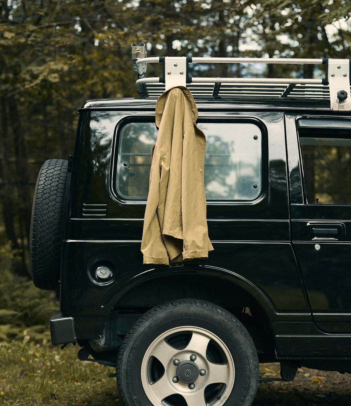The Best Made Co. Possibilities Jacket Tan hangs on the roof rack of a black SUV parked outdoors, framed by trees and foliage in the background.