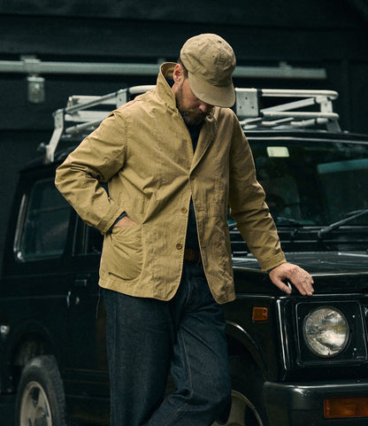 A man wearing the Best Made Co. Possibilities Jacket in tan and a cap leans against a black SUV, looking down with one hand in his pocket. The background is dark, featuring a metal structure on the car’s roof.
