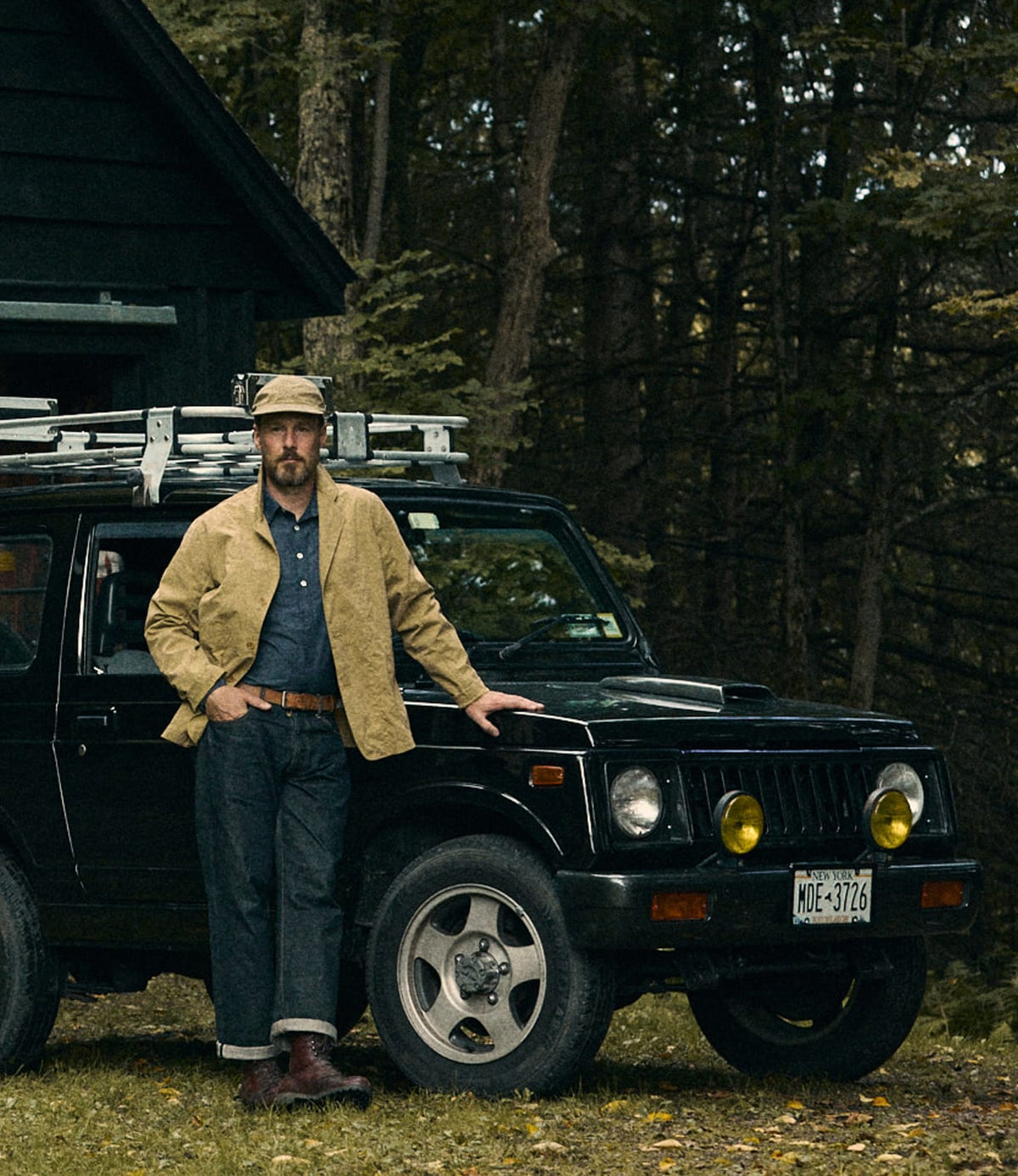 A man wearing the Best Made Co. Possibilities Jacket in tan and a hat leans against a black SUV equipped with a ladder on the roof, parked on grass beside a dark wooden building and trees.