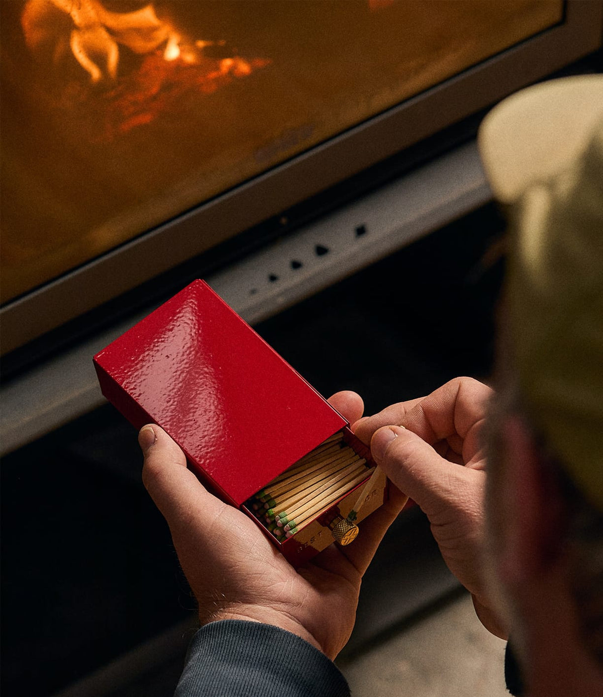 Hands holding an open red metal Sliding Match Safe Red filled with strike-anywhere matches in front of a fireplace.