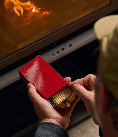 Hands holding an open red metal Sliding Match Safe Red filled with strike-anywhere matches in front of a fireplace.