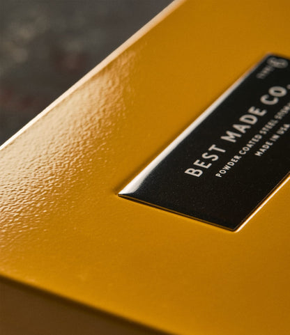 Close-up of a textured yellow durable steel box, the Type 6 Strongbox Yellow by Best Made Co. Featuring a shiny, slightly raised black label with the brand name, it exudes a premium feel and is ideal for secure storage.