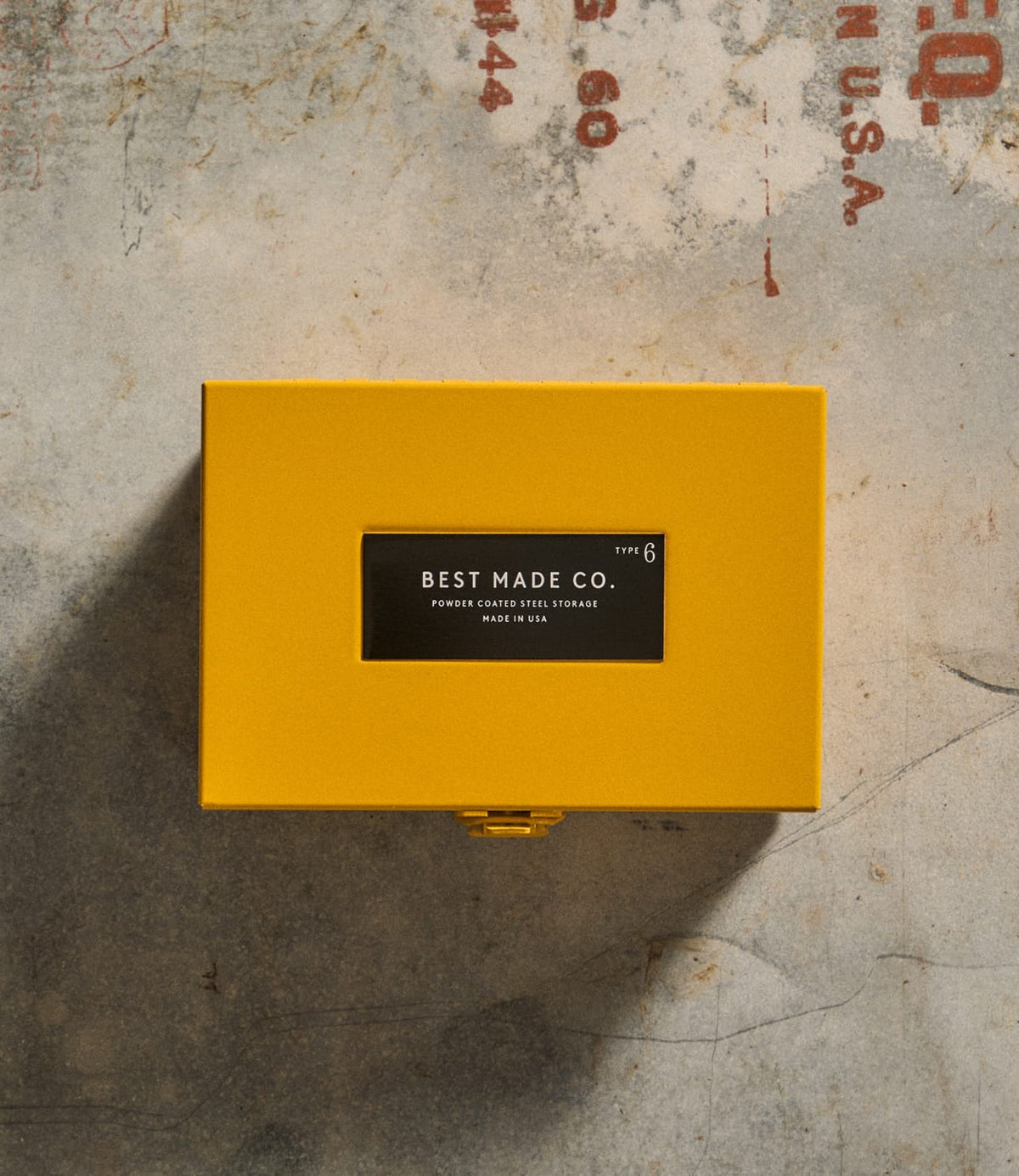 A durable steel box labeled Best Made Co. Type 6 Strongbox Yellow sits on a textured concrete surface with red stenciled text partially visible, offering safe storage for your essentials.