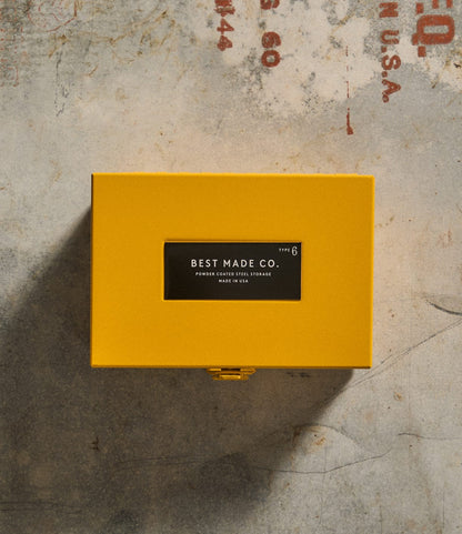 A durable steel box labeled Best Made Co. Type 6 Strongbox Yellow sits on a textured concrete surface with red stenciled text partially visible, offering safe storage for your essentials.