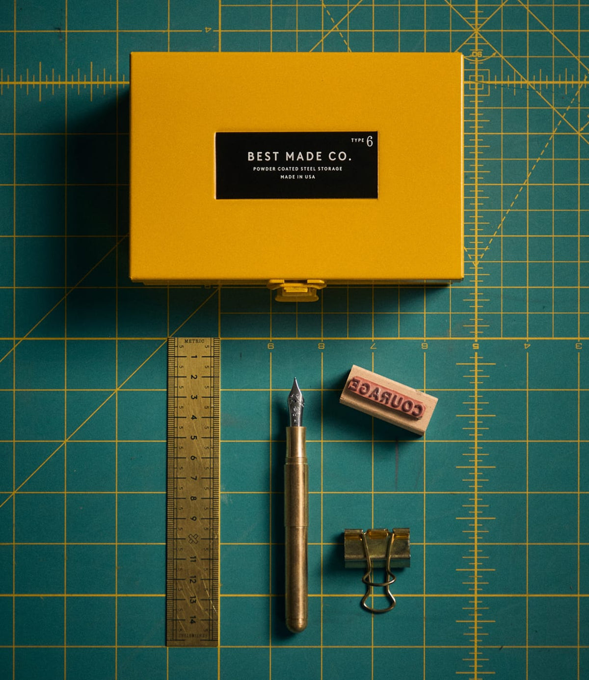 Best Made Co. Type 6 Strongbox Yellow sits atop a green cutting mat, accompanied by a metal pen, gold ruler, brown eraser with “Courage” text, and a gold binder clip—all neatly arranged for an organized flat lay workspace aesthetic and secure storage.