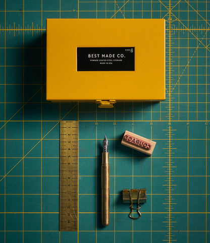 Best Made Co. Type 6 Strongbox Yellow sits atop a green cutting mat, accompanied by a metal pen, gold ruler, brown eraser with “Courage” text, and a gold binder clip—all neatly arranged for an organized flat lay workspace aesthetic and secure storage.