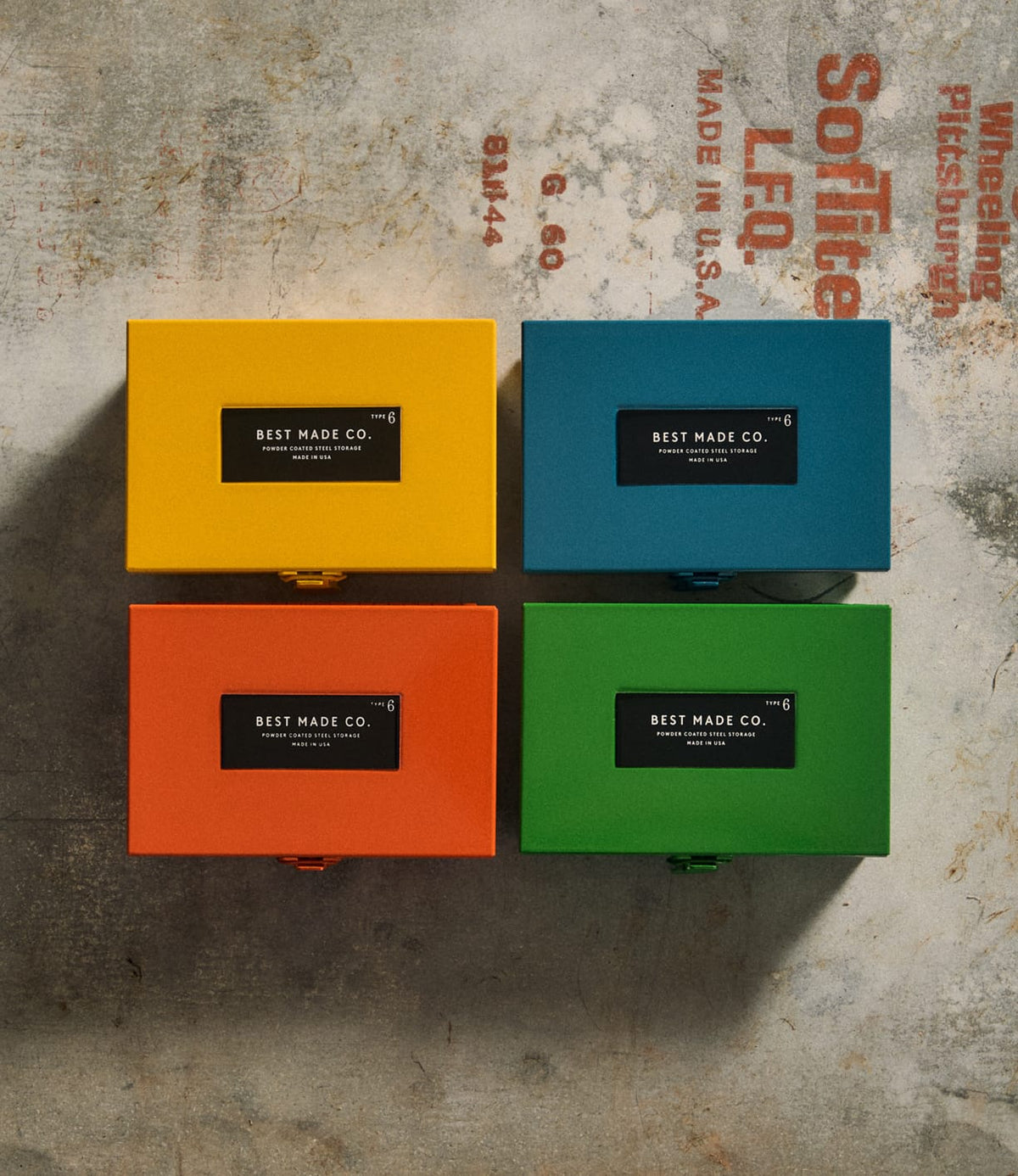 Four vibrant yellow metal strongboxes—each a Type 6 Strongbox Yellow by Best Made Co.—are arranged in a square on a concrete surface adorned with faded industrial text. Each durable steel box prominently features a black label reading “Best Made Co.,” providing secure storage for your essentials.