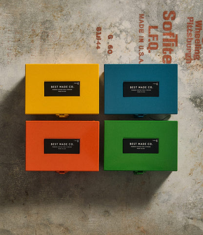 Four vibrant yellow metal strongboxes—each a Type 6 Strongbox Yellow by Best Made Co.—are arranged in a square on a concrete surface adorned with faded industrial text. Each durable steel box prominently features a black label reading “Best Made Co.,” providing secure storage for your essentials.