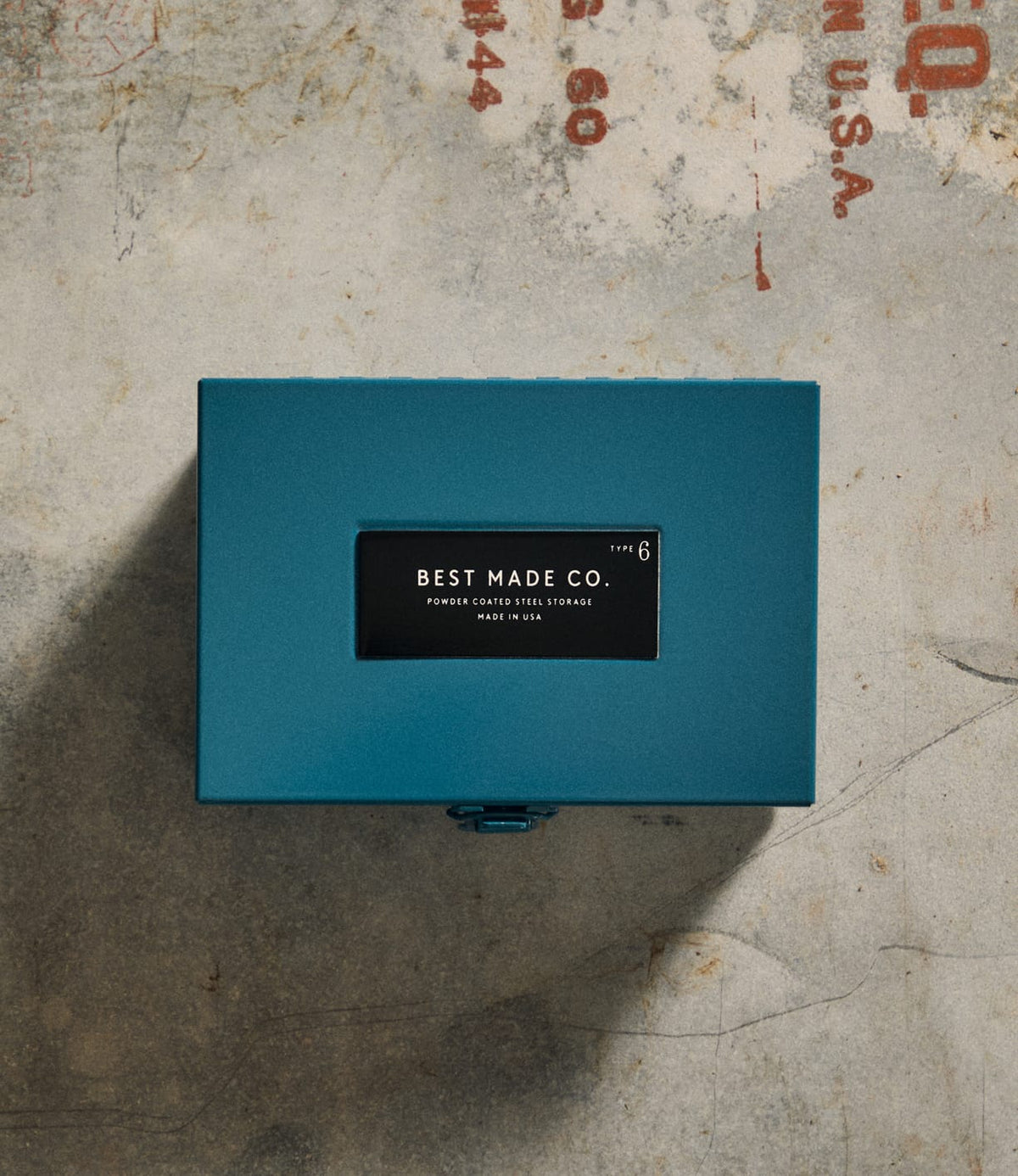 A limited edition Type 6 Strongbox Blue from Best Made Co., featuring a striking teal metal finish and a black Best Made Co. label, sits atop a worn concrete surface with faded red lettering, exemplifying the exceptional quality and craftsmanship of Best Made strongboxes.