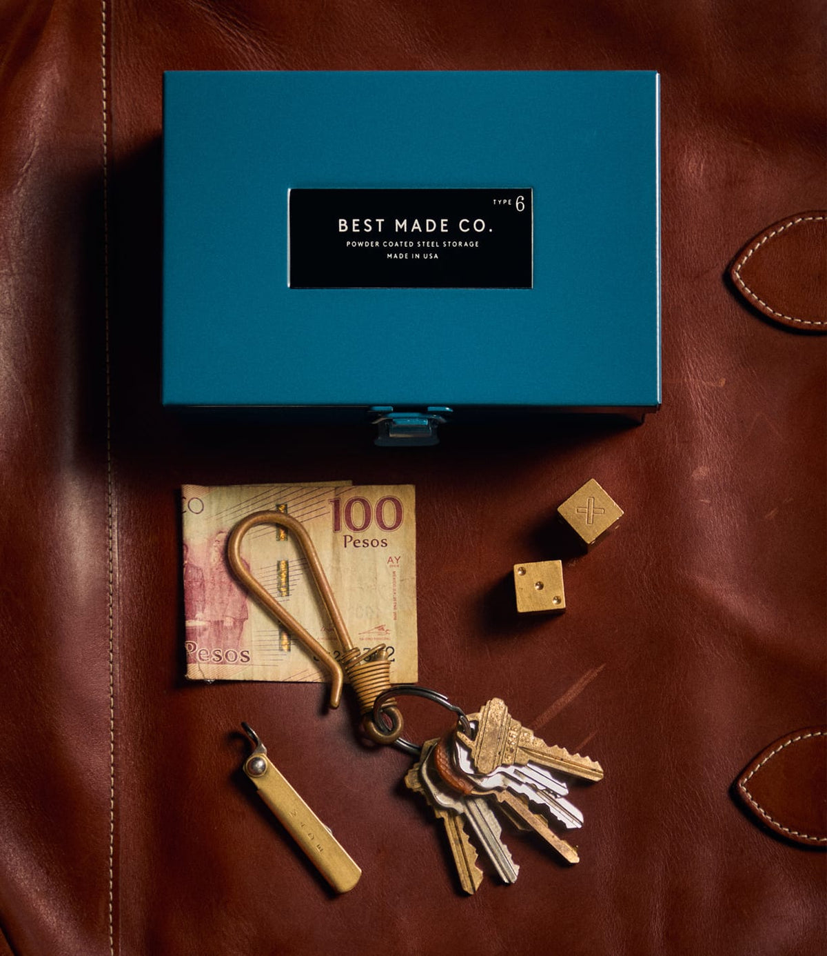 A limited edition Best Made Co. Type 6 Strongbox Blue, a 100 pesos bill, a gold carabiner holding several keys, a small pocket tool, and two brass dice rest on a brown leather surface.
