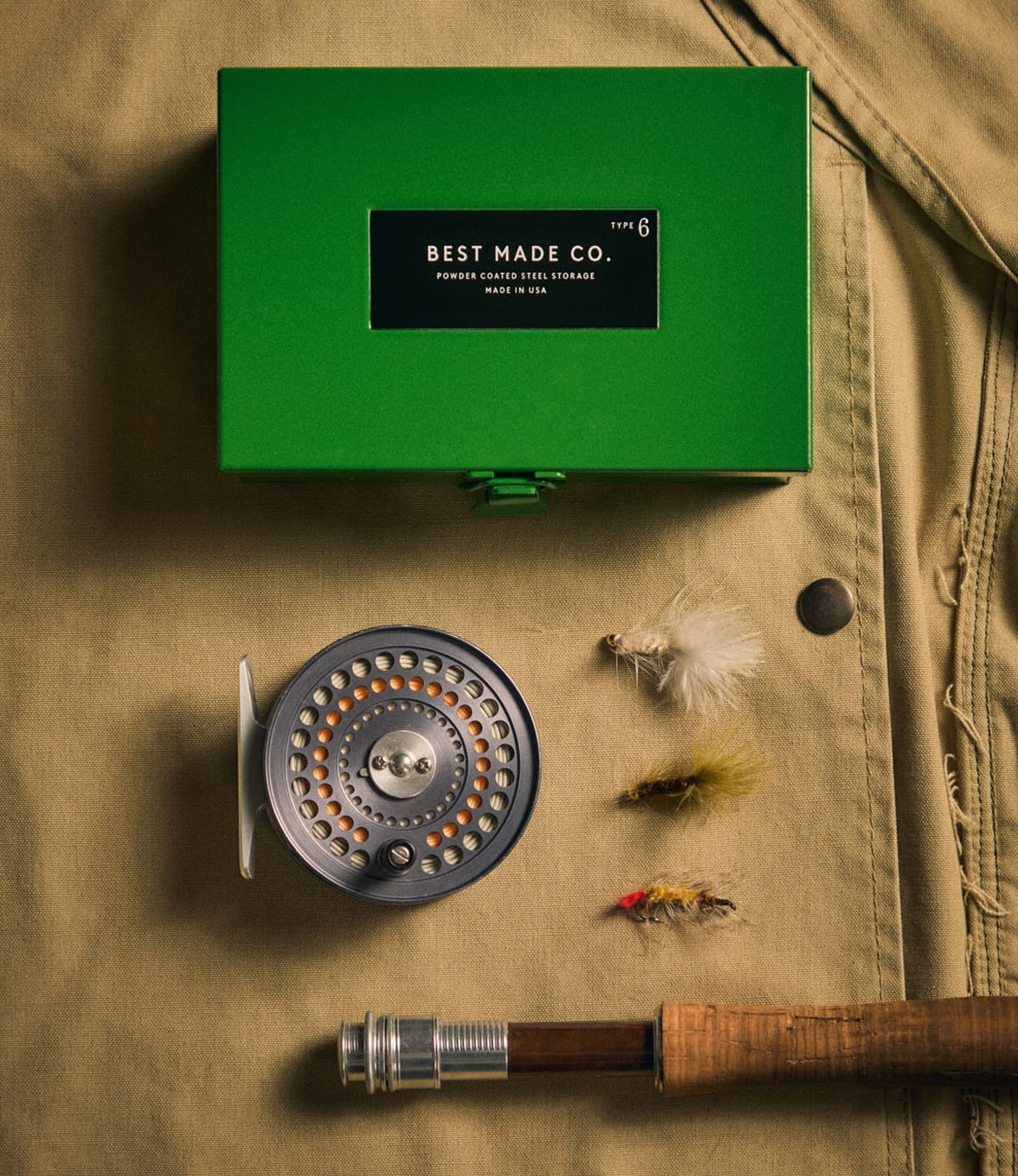 A durable steel Type 6 Strongbox Green by Best Made Co., a fishing reel, three fishing flies, and part of a fishing rod are arranged on a tan fabric background, suggesting secure storage for your prized angling gear.