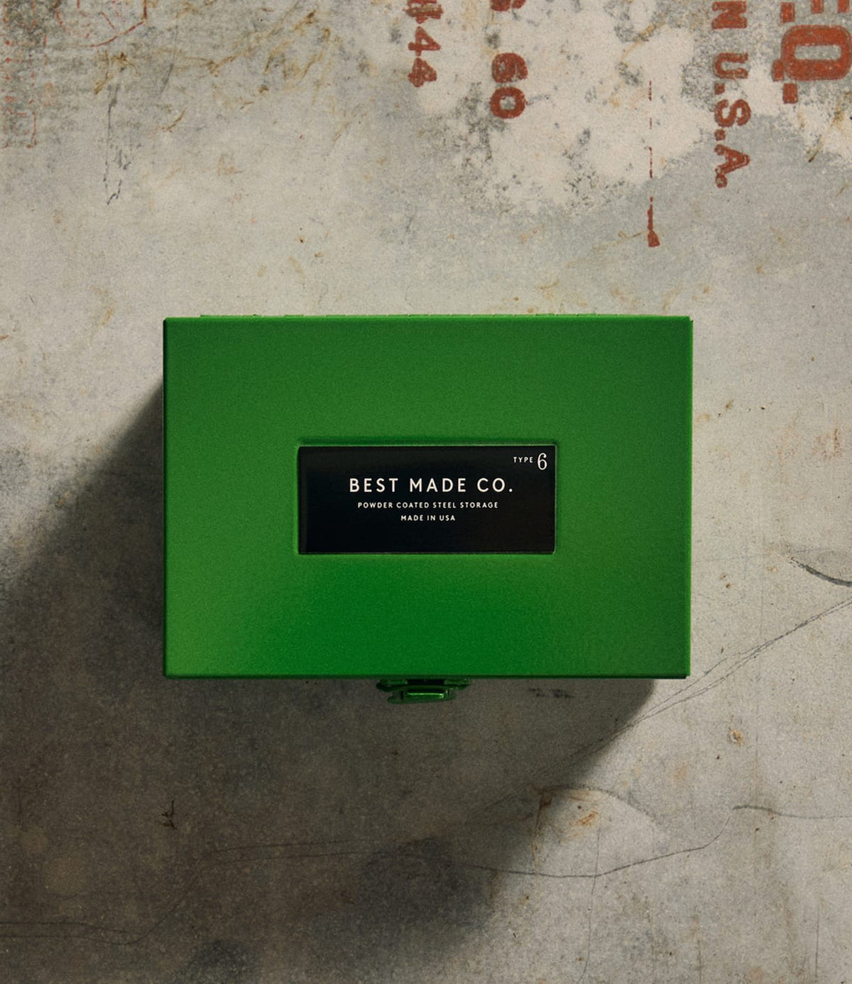 A durable steel Type 6 Strongbox Green from Best Made Co. rests on a worn gray concrete surface with faint red markings, offering secure storage in the spirit of classic strongboxes.