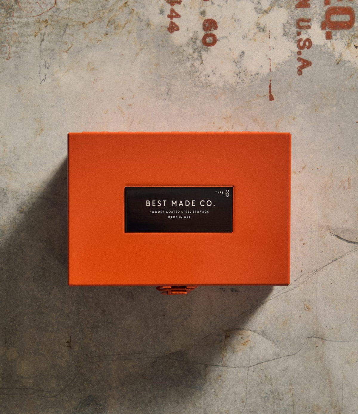 The Type 6 Strongbox Orange by Best Made Co. rests on a textured concrete surface. This durable steel box features a vivid orange finish, a black and white label displaying the brand name and product details, and a prominent front latch—reflecting the exceptional craftsmanship characteristic of all Best Made Co. strongboxes.