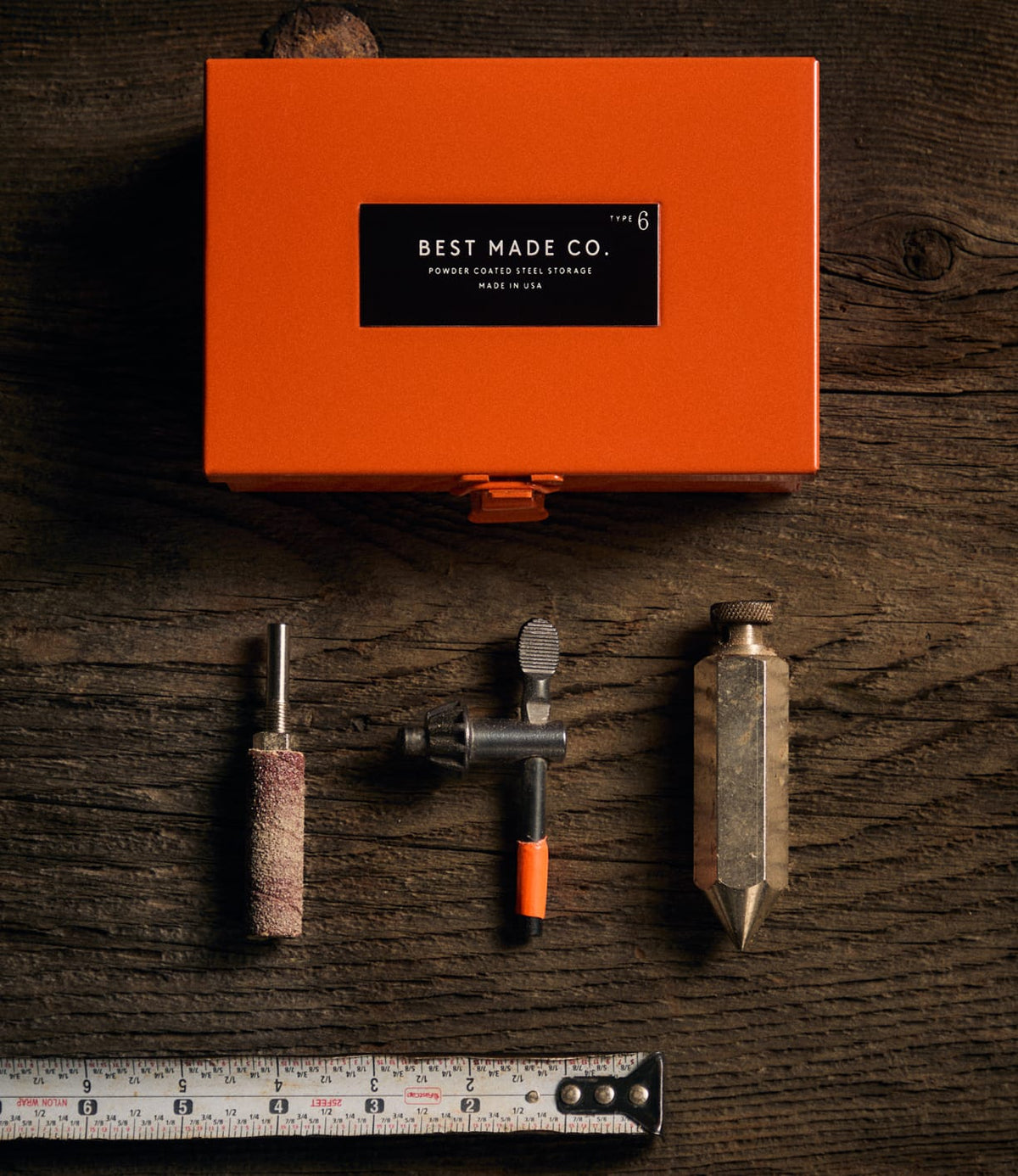 A Best Made Co. Type 6 Strongbox Orange, crafted from durable steel, rests on a wooden surface above three metal tools and a measuring tape, all neatly arranged side by side.