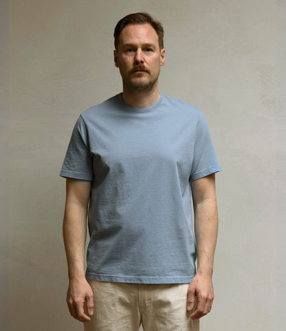 A man with short brown hair and a beard stands against a plain, light wall, wearing Best Made Co.’s Standard T-Shirt in Overcast Blue and off-white pants. He looks directly at the camera with a neutral expression.