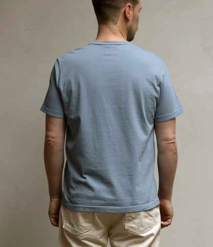 A man faces away from the camera, wearing the Best Made Co. Standard T-Shirt in Overcast Blue with beige pants against a neutral, textured background.