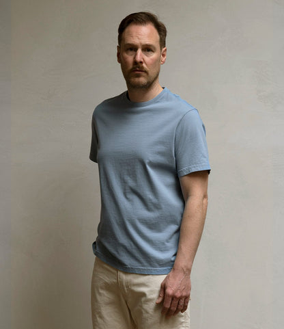A man with short brown hair and a beard stands against a light background wearing the Best Made Co. Standard T-Shirt in Overcast Blue and off-white pants, looking forward with a neutral expression.