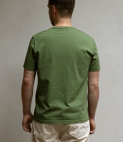 A person faces away from the camera, wearing Best Made Co.’s Standard T-Shirt in Olive Drab and light pants, set against a plain neutral background.