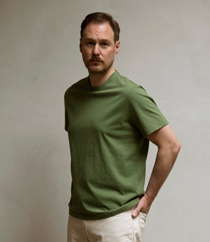 A man with short hair and a beard stands against a plain light-colored wall, wearing the Best Made Co. Standard T-Shirt in Olive Drab and light beige pants, looking at the camera with a neutral expression and one hand behind his back.
