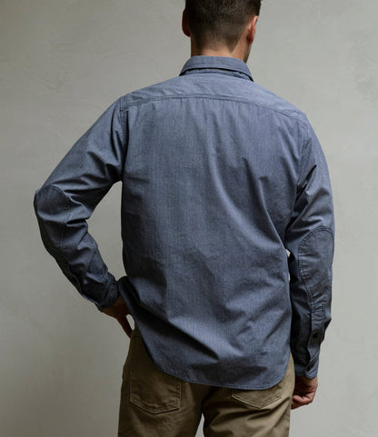 A person stands with their back to the camera, wearing the Best Made Co. Indigo Stripe Workshirt made from Portuguese cotton and tan pants, one hand on their hip against a plain, light-colored wall.