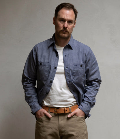 A man with short brown hair and a trimmed beard stands against a plain light wall, wearing the Best Made Co. Indigo Stripe Workshirt over a white T-shirt, beige pants, and a tan belt, with his hands in his pockets.