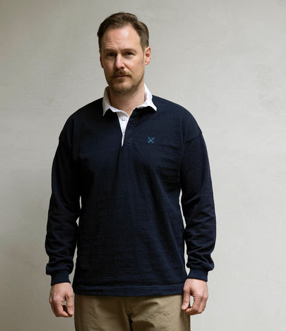 A man with short hair and light facial hair stands against a plain light wall, wearing the Best Made Co. McArthur Rugby Jersey in navy blue over a white collared shirt and tan pants, looking directly at the camera with a neutral expression.