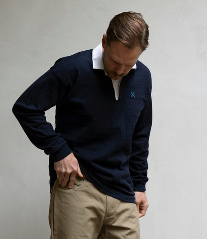 A man wears the Best Made Co. McArthur Rugby Jersey—navy heavyweight cotton with a white collar—and khaki pants, looking down with one hand in his pocket against a plain light-colored wall.