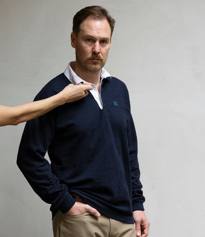 A man wears the Best Made Co. McArthur Rugby Jersey in navy with khaki pants, standing against a plain light background while someone off-frame adjusts his white collar.
